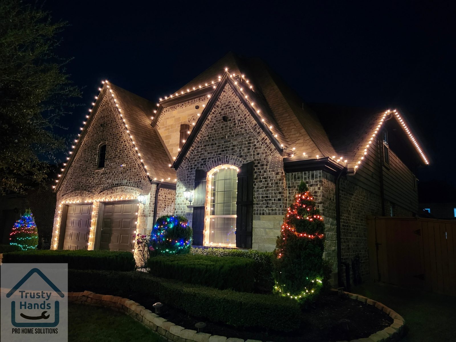 Stone home in Katy, TX with warm white roofline lighting and colorful lit shrubs