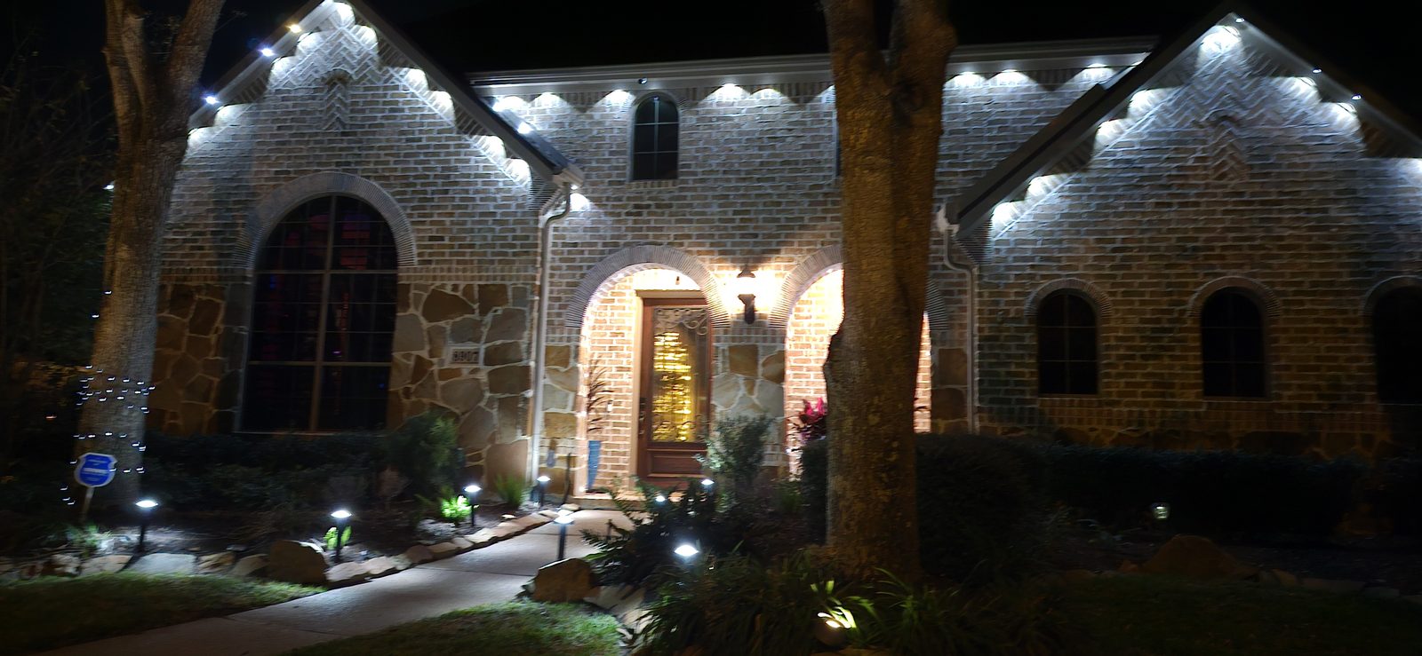Warm white permanent roofline and landscape lighting on a stone home entrance in Katy, TX